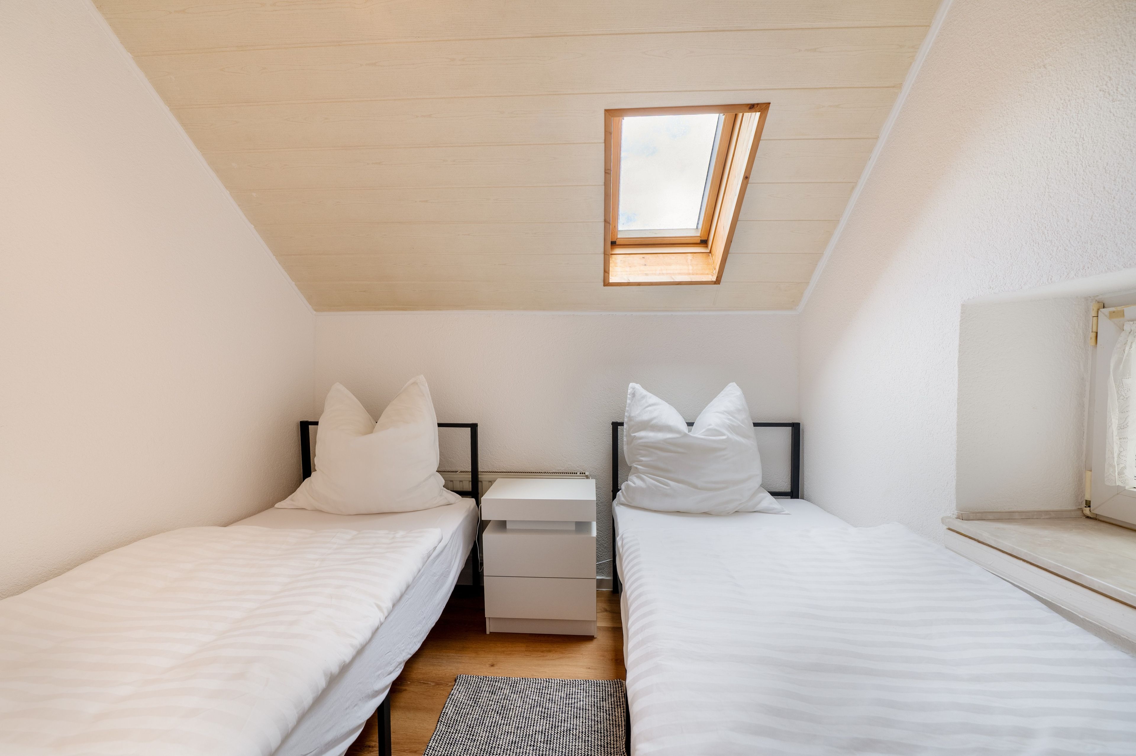 A small, single-room bedroom with two beds, a skylight, and a white wardrobe.