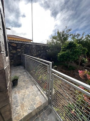Un balcón de baldosas de piedra con baranda de metal, un muro de piedra y vista a un jardín con diversas plantas y un árbol.