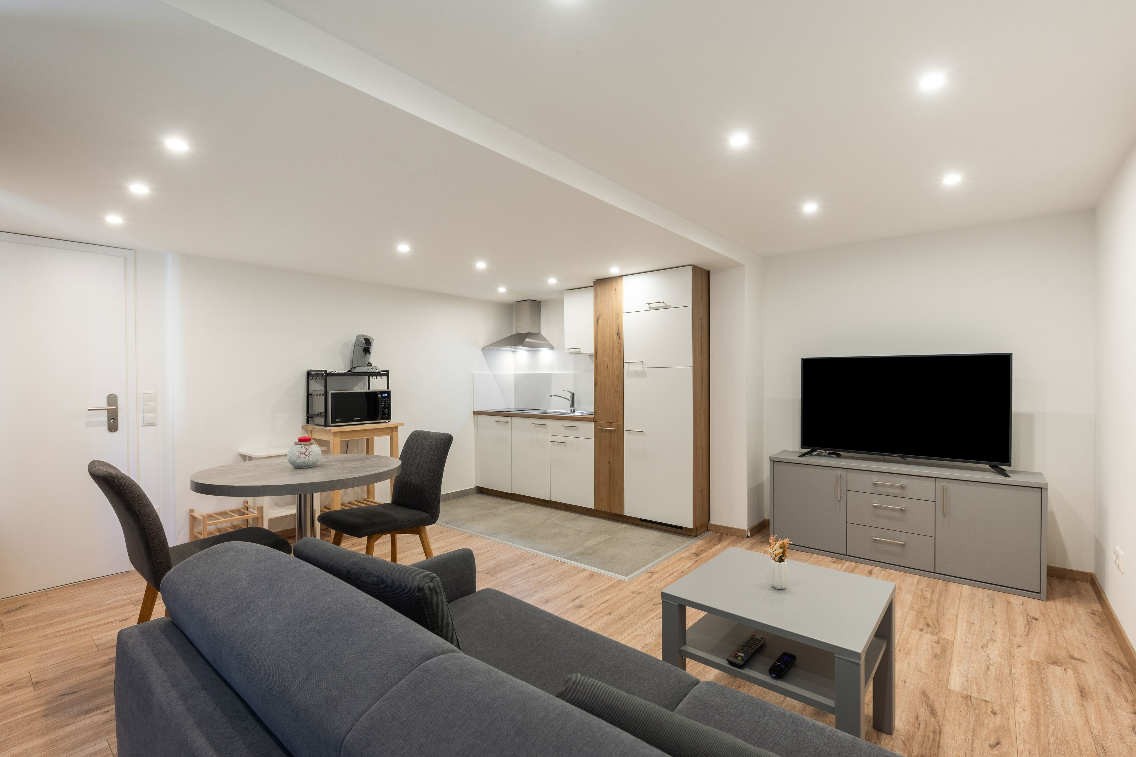 Un salon moderne avec un canapé gris, une petite table ronde et un téléviseur à écran plat posé sur un meuble gris.