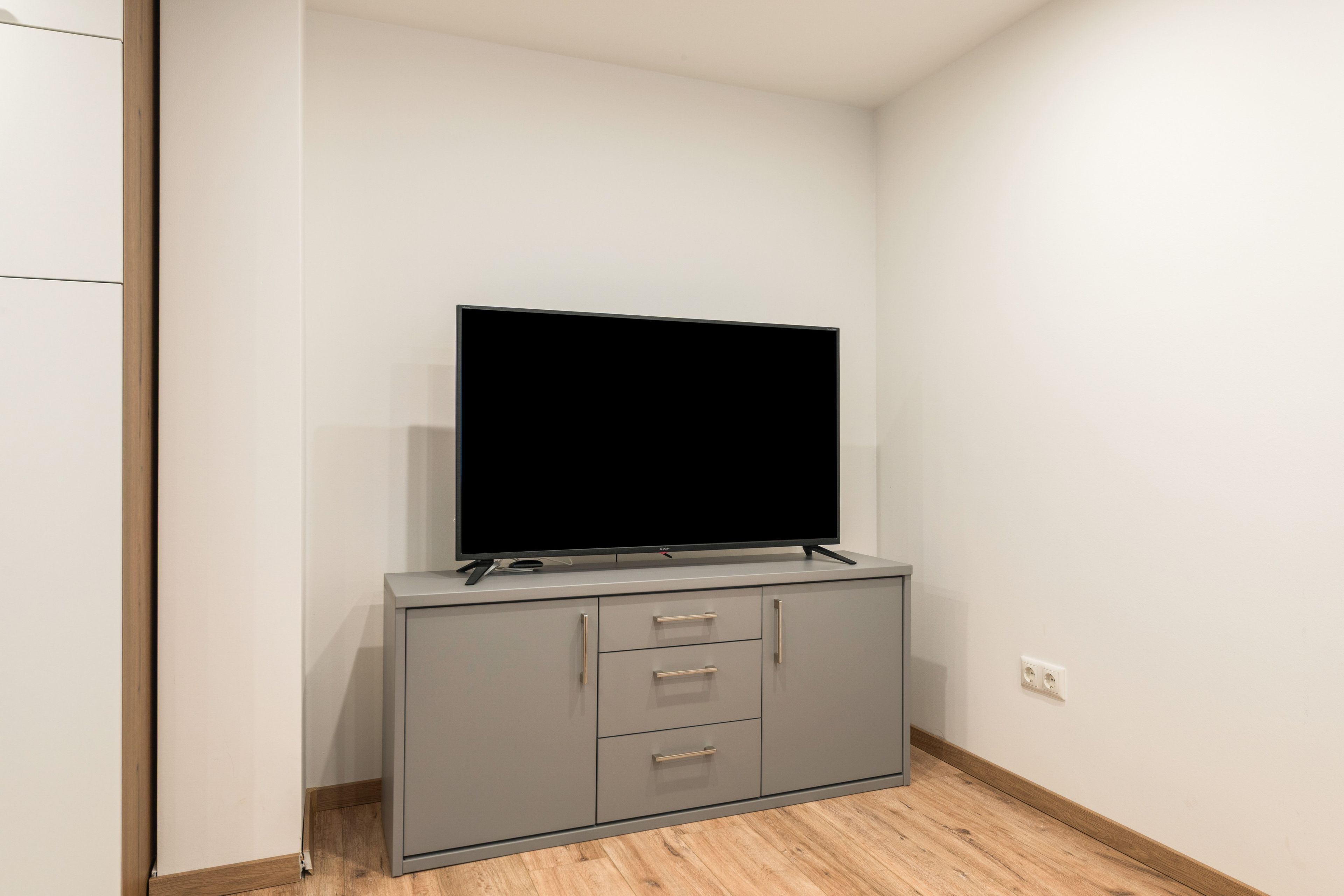Une pièce moderne et minimaliste, dotée d’un téléviseur à écran plat fixé sur un meuble gris, d’un plancher en bois et de murs blancs.