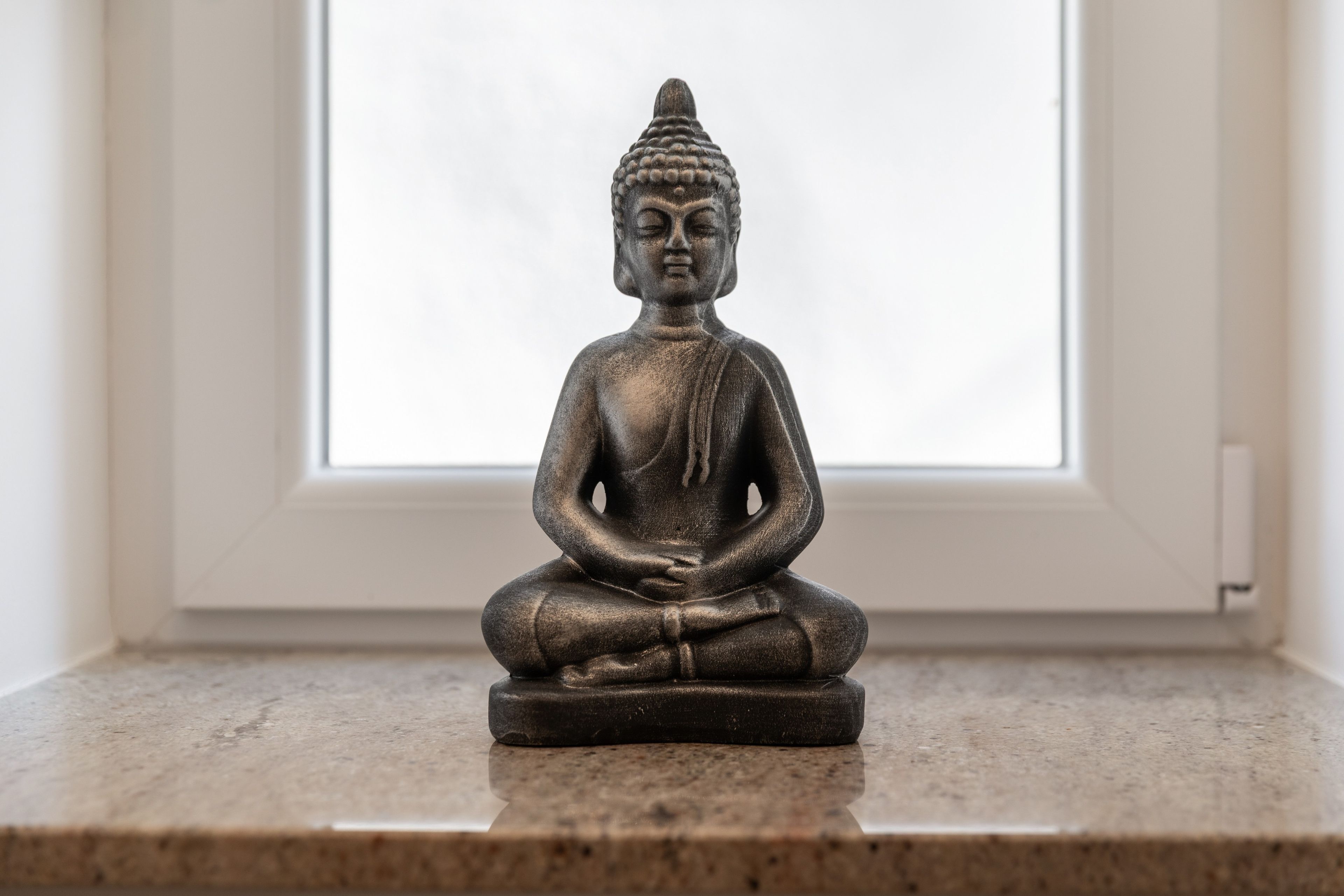 A statue of a seated figure on a pedestal in a room with a window.