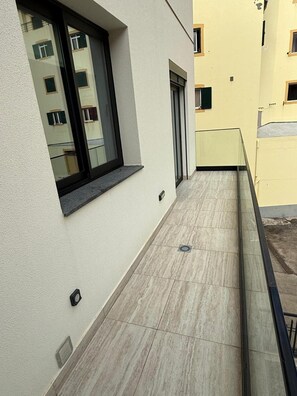 Ein schmaler Außenkorridor mit Fliesenboden, einem Fenster mit schwarzem Rahmen und einer Tür mit grünem Rollo.