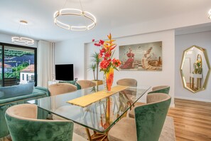 Een moderne eetruimte met een glazen tafel, groene bekleden stoelen, een vaas met bloemen en een groot schilderij aan de muur.