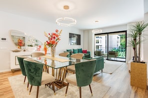 Een moderne woonkamer met een glazen eettafel, groene fluweelstoelen en een grote potplant.