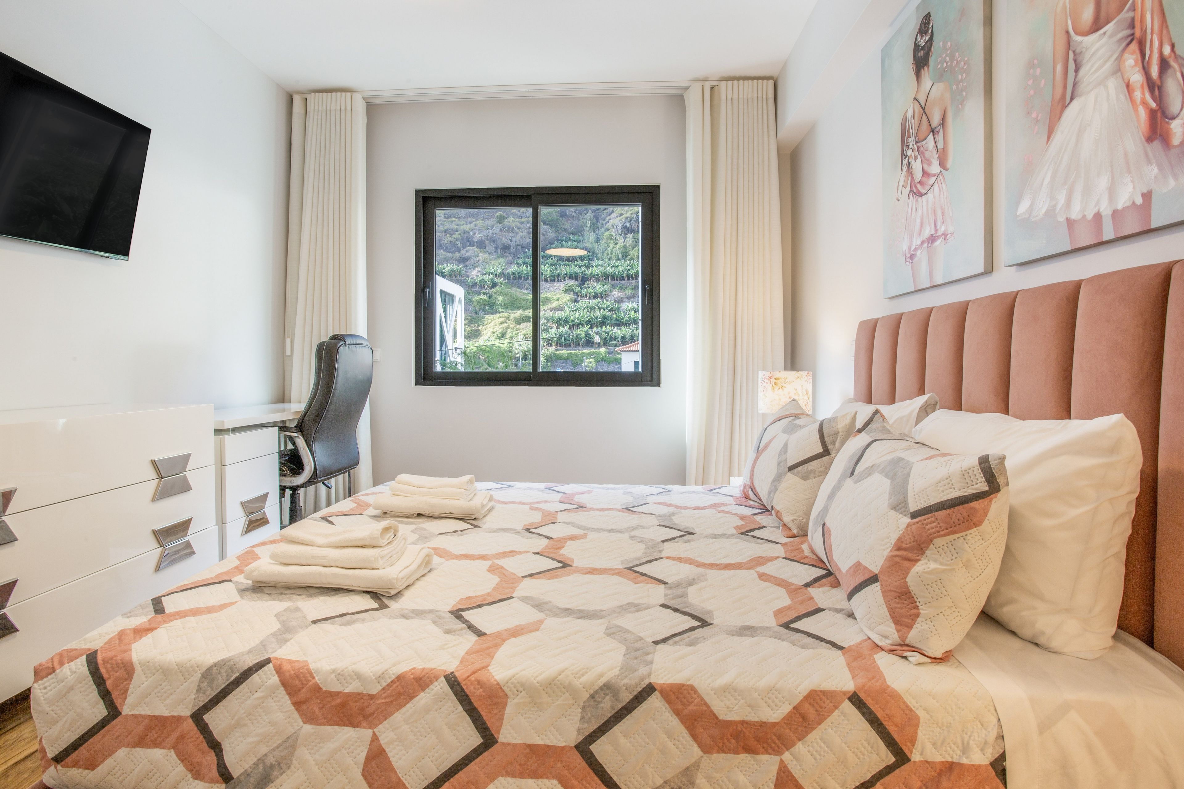 A hotel room with a bed, a desk, a chair, and a window with a view of greenery.