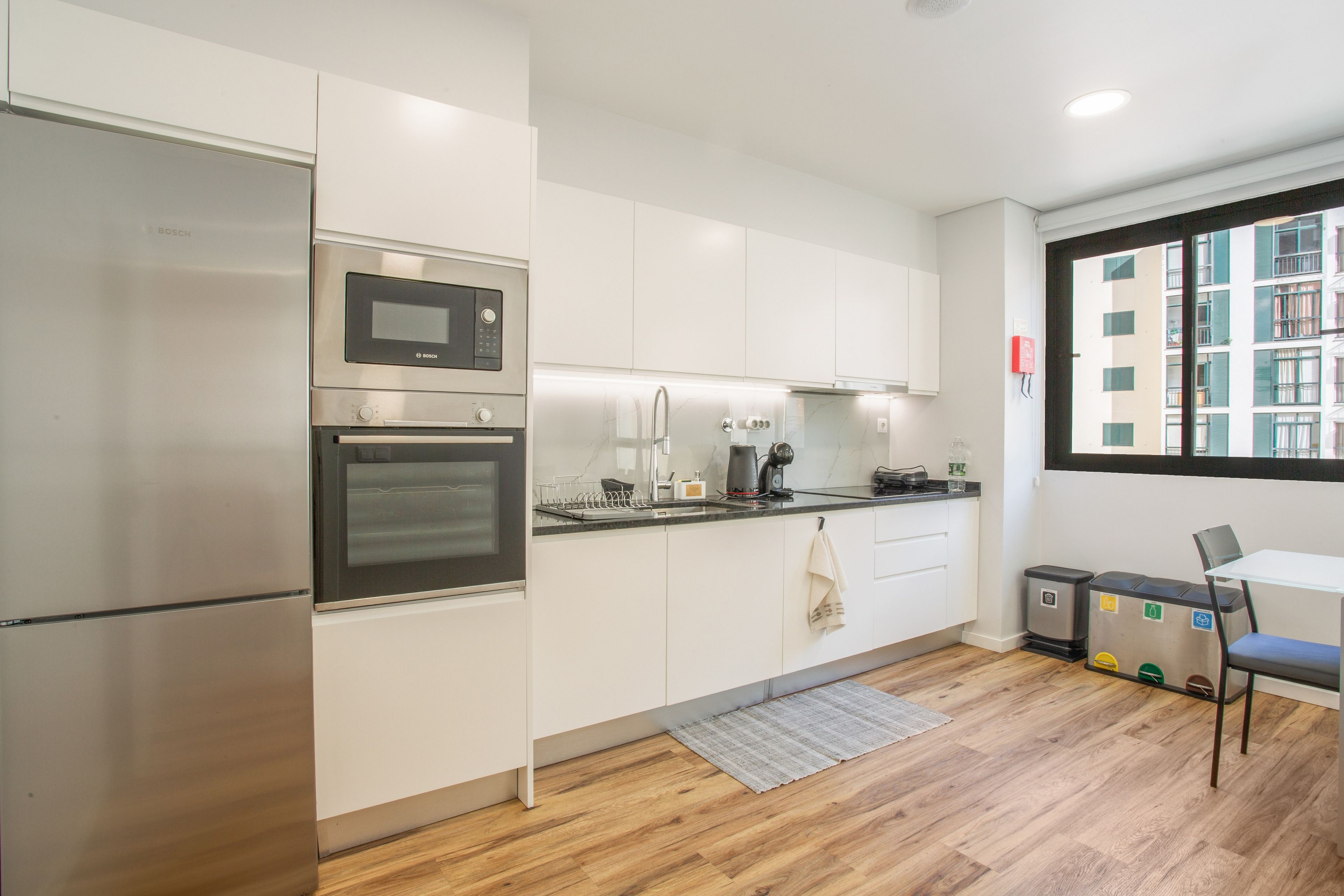 A modern kitchen with stainless steel appliances, a built-in oven, a microwave, and a coffee machine. There is a small dining area with a table and chairs, and a view of a building through a window.