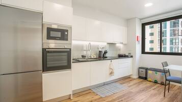 A modern kitchen with stainless steel appliances, a built-in oven, a microwave, and a coffee machine. There is a small dining area with a table and chairs, and a view of a building through a window.