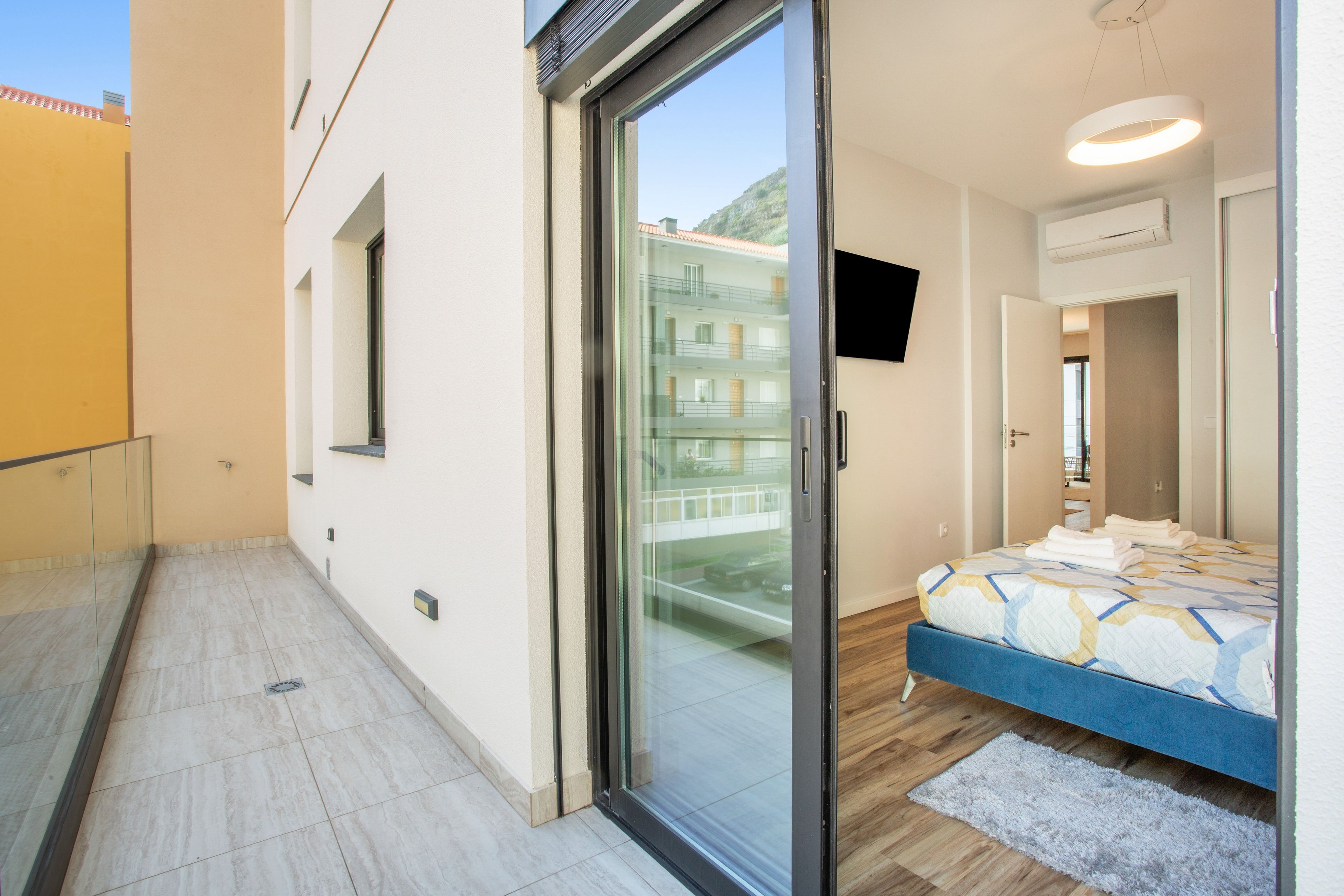 Un dormitorio moderno con un vidrio que da a un balcón, una cama con cubrecama estampado y un televisor montado en la pared.