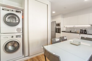 Een moderne keuken met inbouwapparatuur, een eethoek met een glazen tafel, en een wasruimte met gestapelde wasmachines.