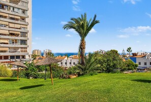 A grassy area with a palm tree, a thatched umbrella, and a building with balconies.