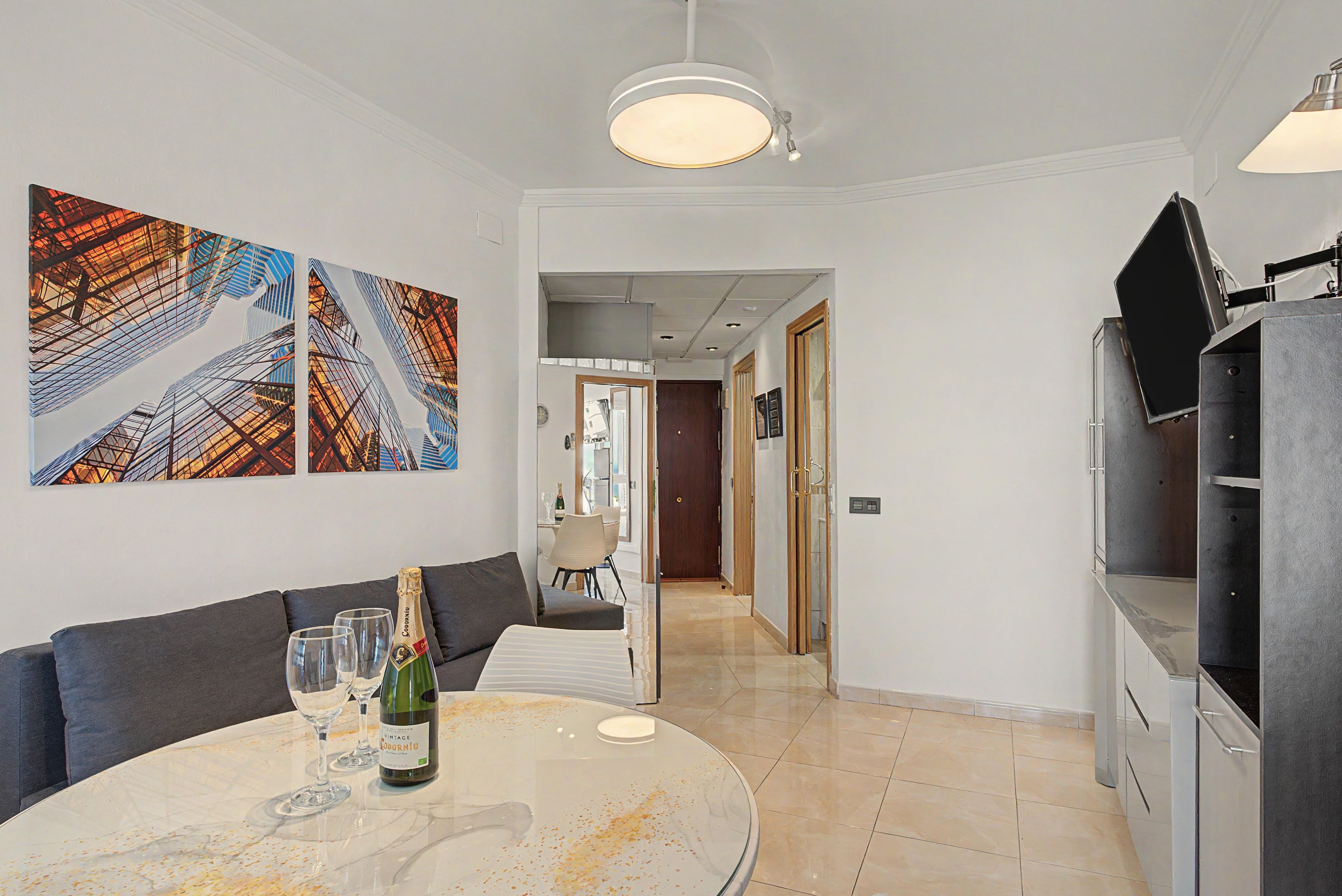 A modern living room with a round table, a sofa, and a TV stand. There are two abstract paintings on the wall and a ceiling light fixture.