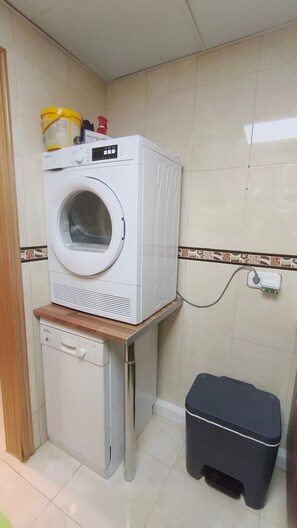 A compact laundry area with a washing machine and a dryer stacked on a wooden platform, a small trash bin, and a power outlet on the wall. - La Colina Apartment with Sea Views, Wi-Fi, and Air Conditioning (Torremolinos)