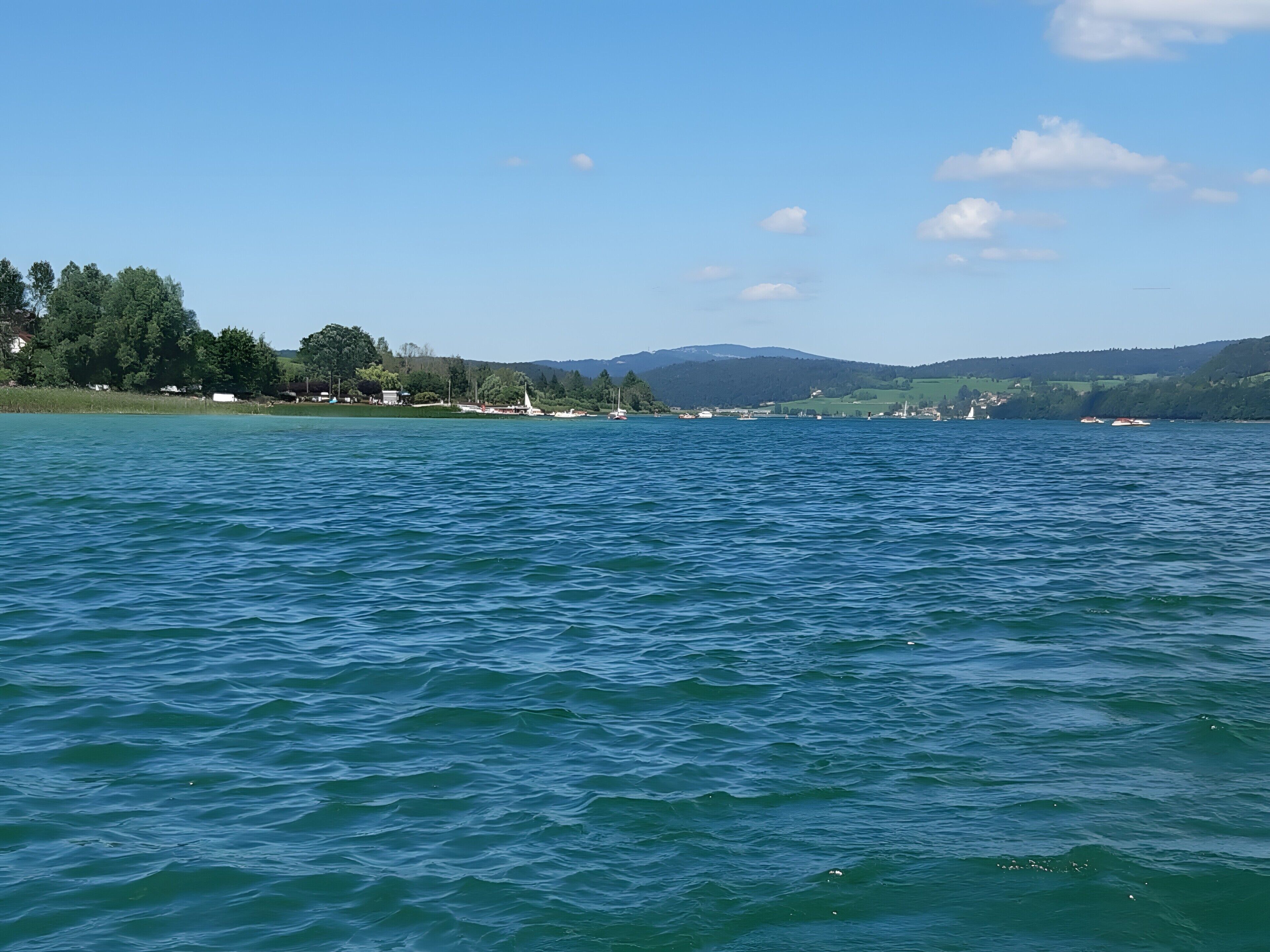 Un lac paisible où l’on aperçoit des bateaux, un phare et, au loin, un paysage verdoyant.