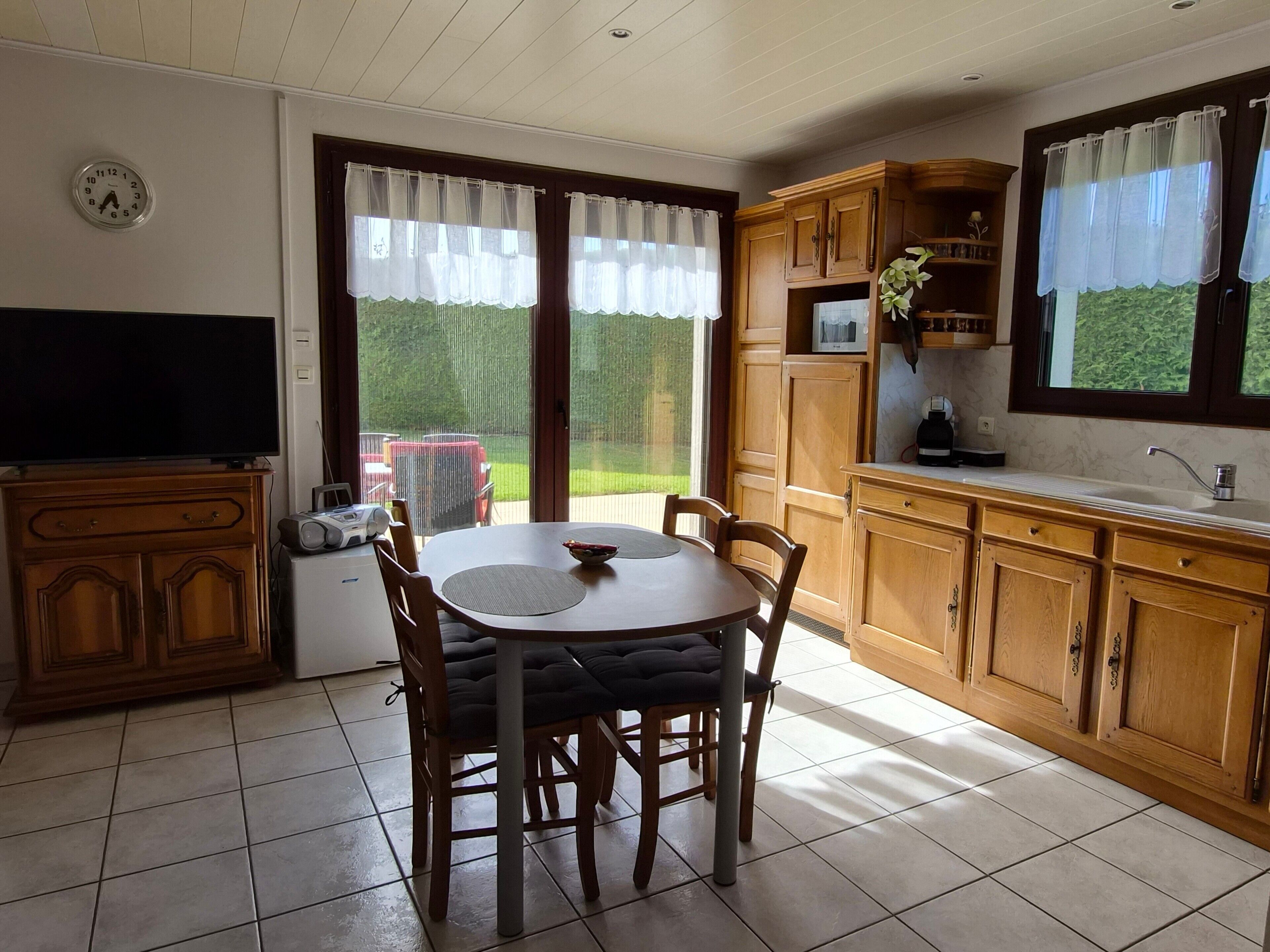 Ein Essbereich mit Tisch und Stühlen, ein Fernseher auf einer Kommode sowie eine Küche mit Holzausbau und einem Fenster.