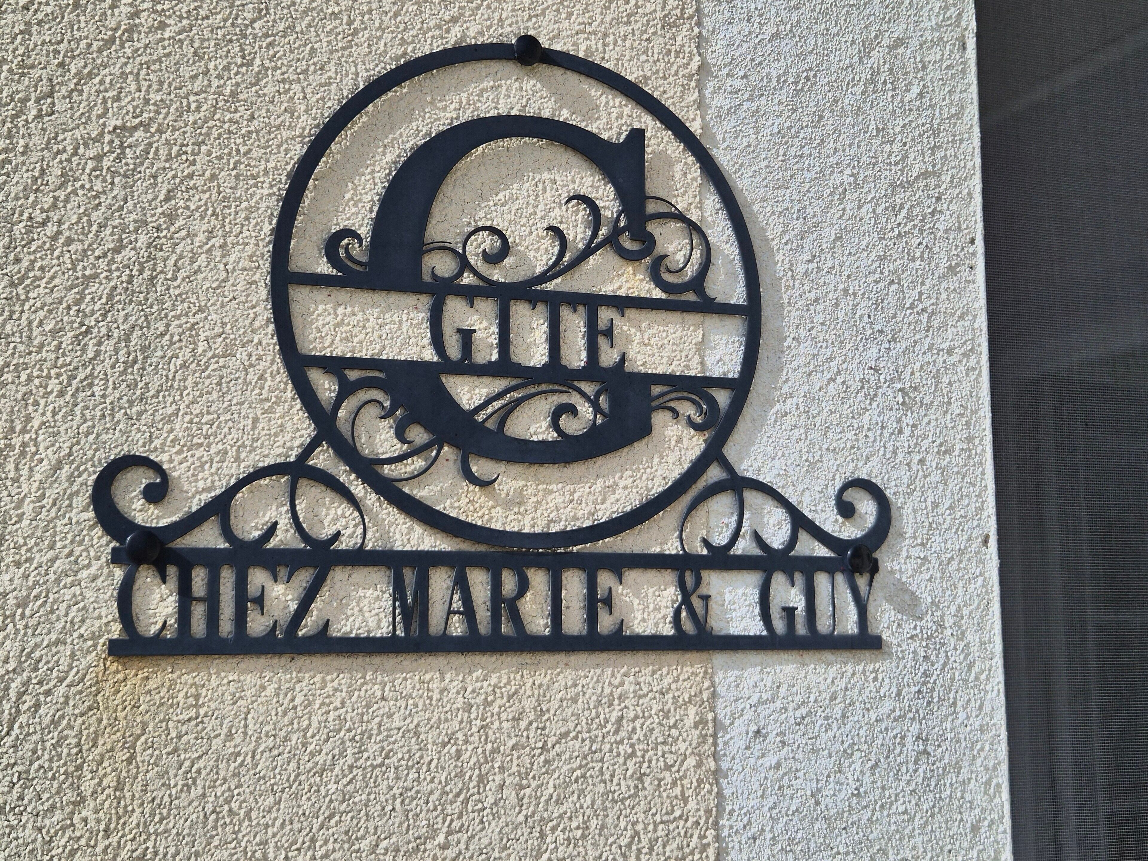 Een metalen bord met de tekst 'CHEZ MARIE & GUN' op een structuurrijke muur.