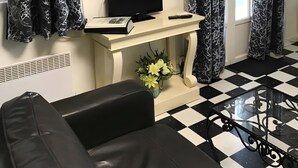 A living room with a black leather sofa, a glass coffee table, a flat-screen TV, a small white console table with a book and remote, and a window with curtains.