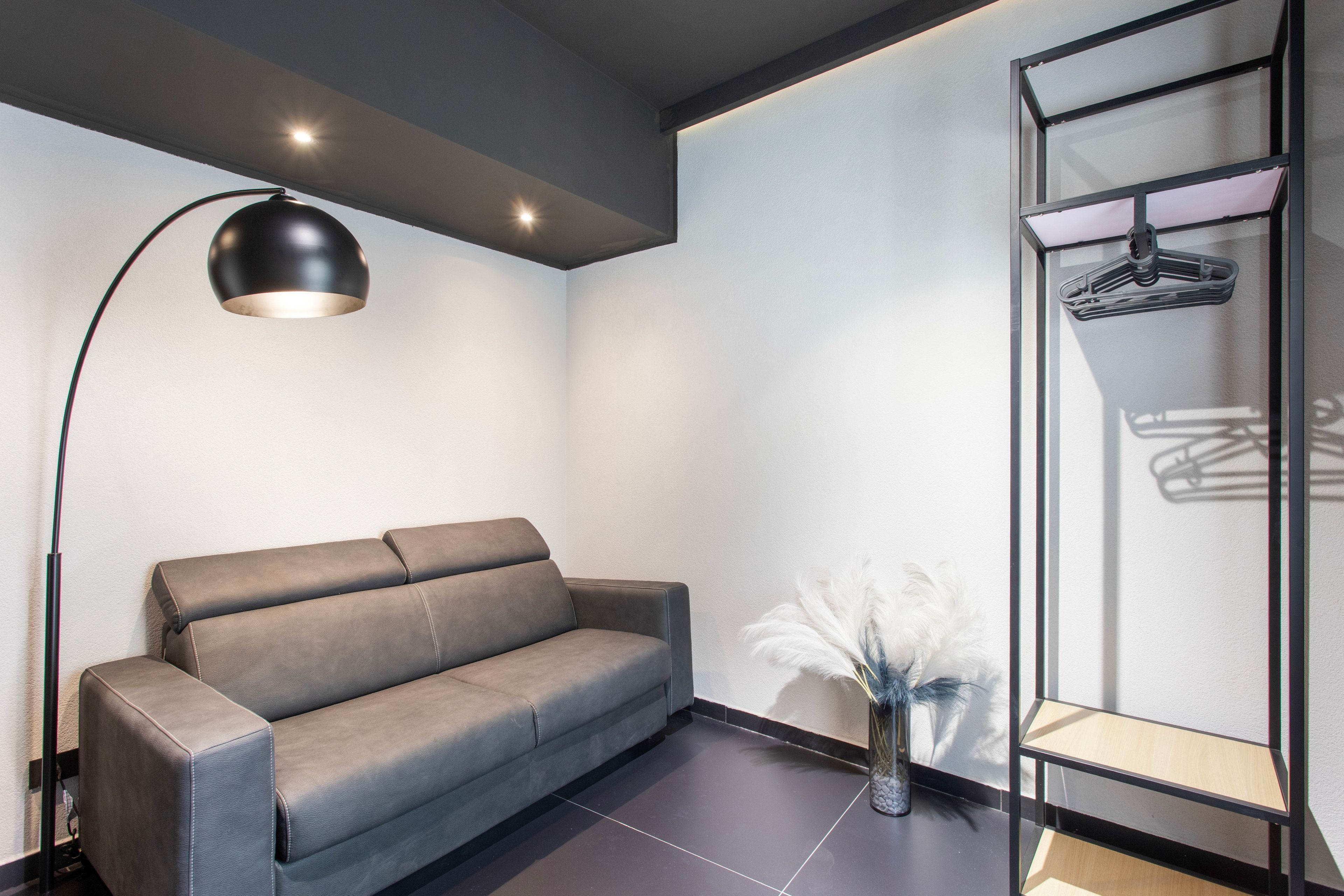 Ein modernes Zimmer mit einem grauen Sofa, einer Stehlampe, einem Glastrennschrank mit einem aufgehängten Kleidungsstück und einer Vase mit weißen Federn.