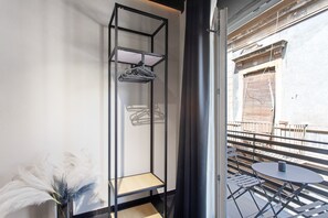 Un balcón con una mesa y sillas, un estante metálico con una percha, y vista a un edificio con ventana amurada.
