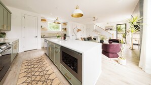 A modern kitchen with a central island, pendant lighting, and a view to a living area with a staircase. - Nashville Rooftop Home with Hot Tub & Skyline Views (Nashville)