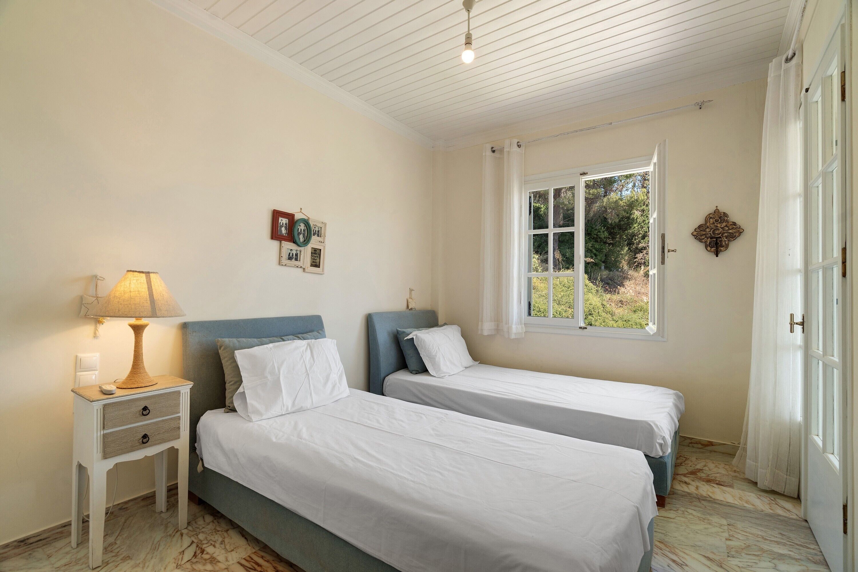 A room with two single beds, a nightstand with a lamp, a window with curtains, and a wall-mounted picture.