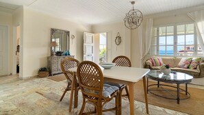 A dining area with a white table, four wicker chairs, a chandelier, and a view of the sea through large windows. - Holiday Home 'Evans Terrace Suite' (Nikiana)