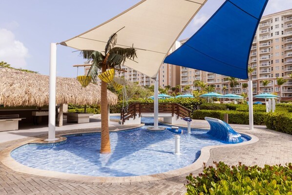 A swimming pool with a wave feature, palm trees, and a thatched roof structure. - Marriott's Aruba Surf Club is on the oceanfront. T (Noord)