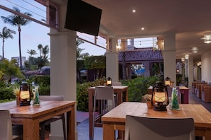 An outdoor dining area with wooden tables and chairs, lanterns, and a view of palm trees and buildings.