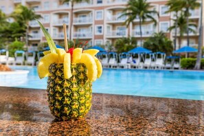 Un cóctel de piña con piscina y hotel al fondo.