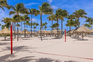 A beach volleyball court with net, surrounded by palm trees and thatched-roof structures.