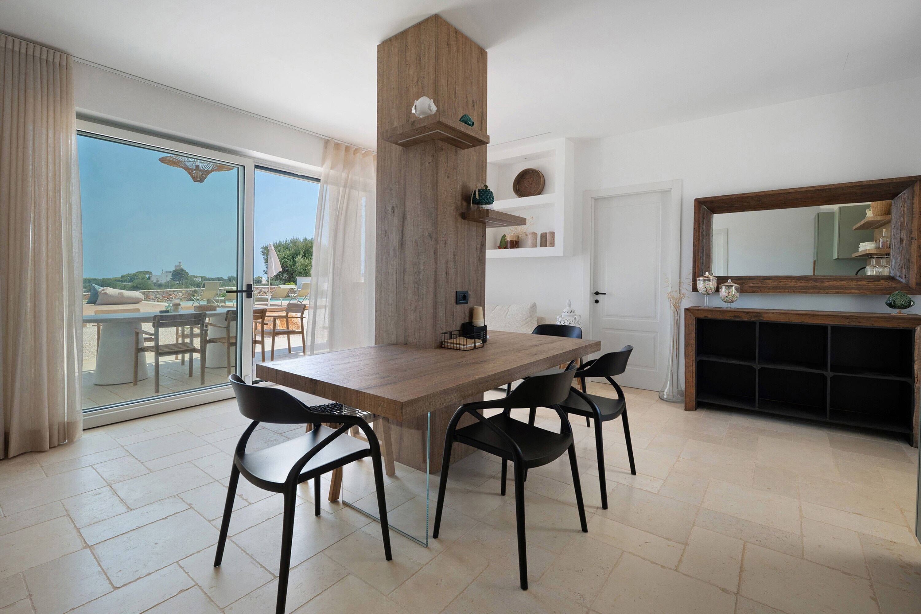 A modern dining area with a wooden table and chairs, a built-in shelf with decorative items, and a view of the outdoors through a glass door.