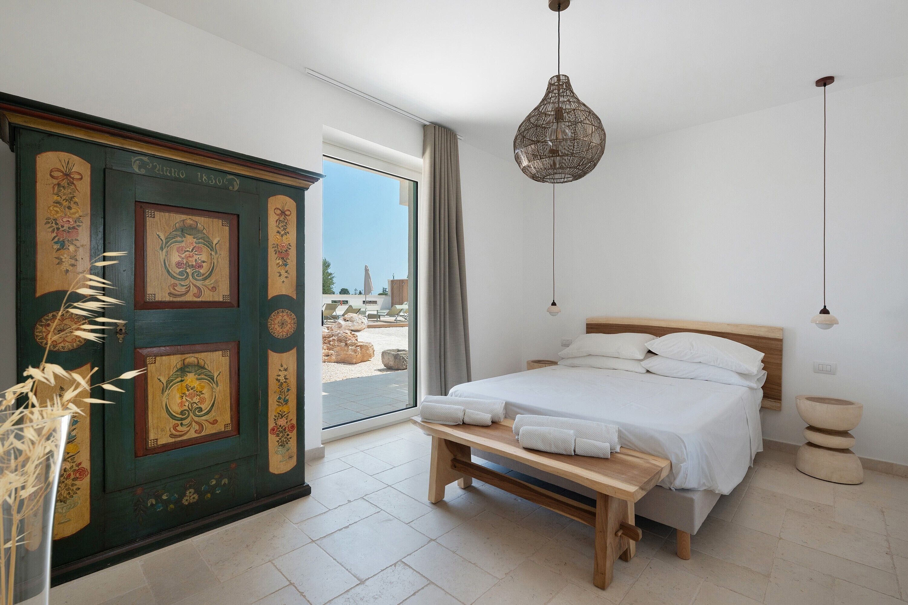 A bedroom with a bed, a bench, a hanging light fixture, and a view of the outside.