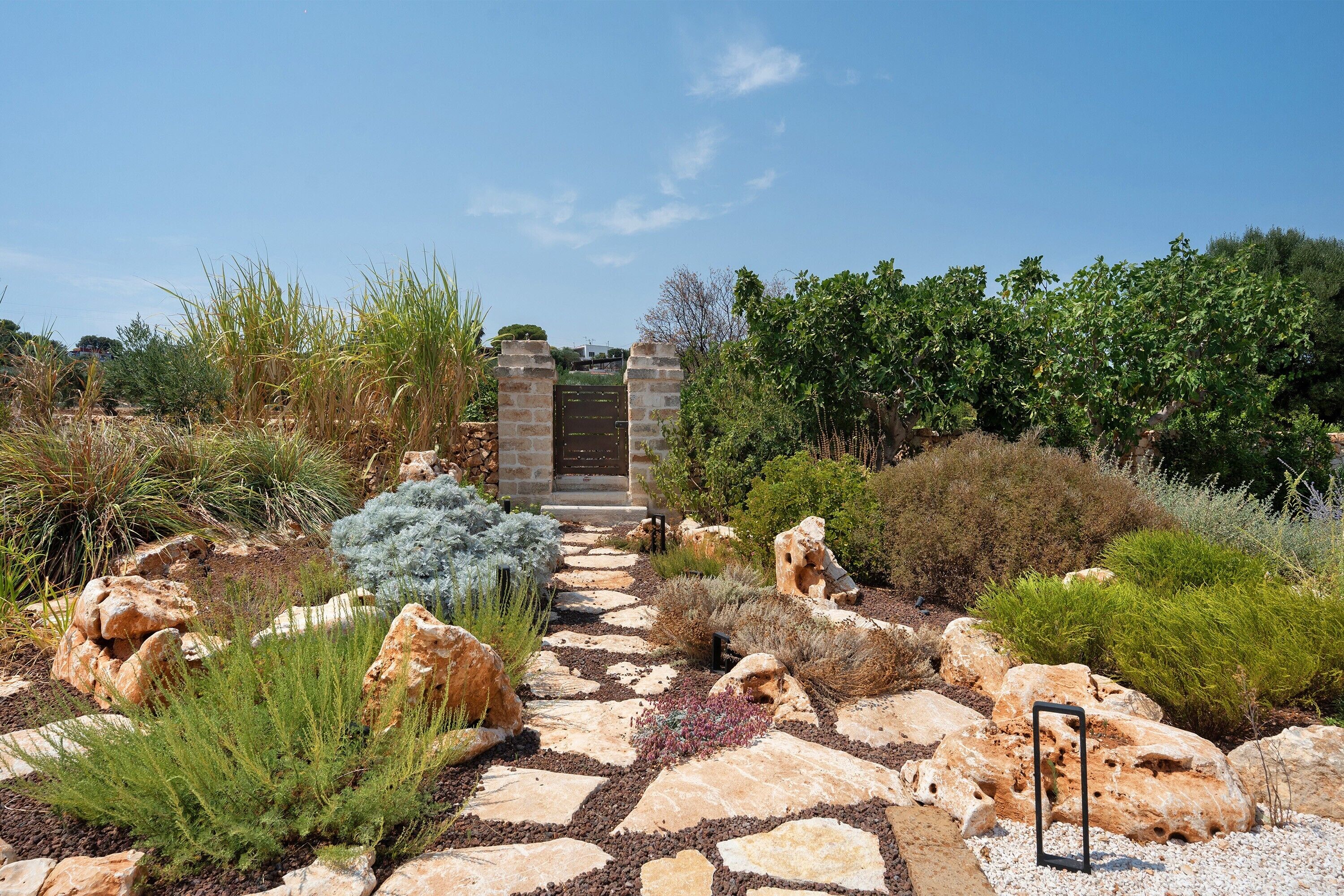 A garden pathway leading to a gated entrance with a stone wall and various shrubs and plants.