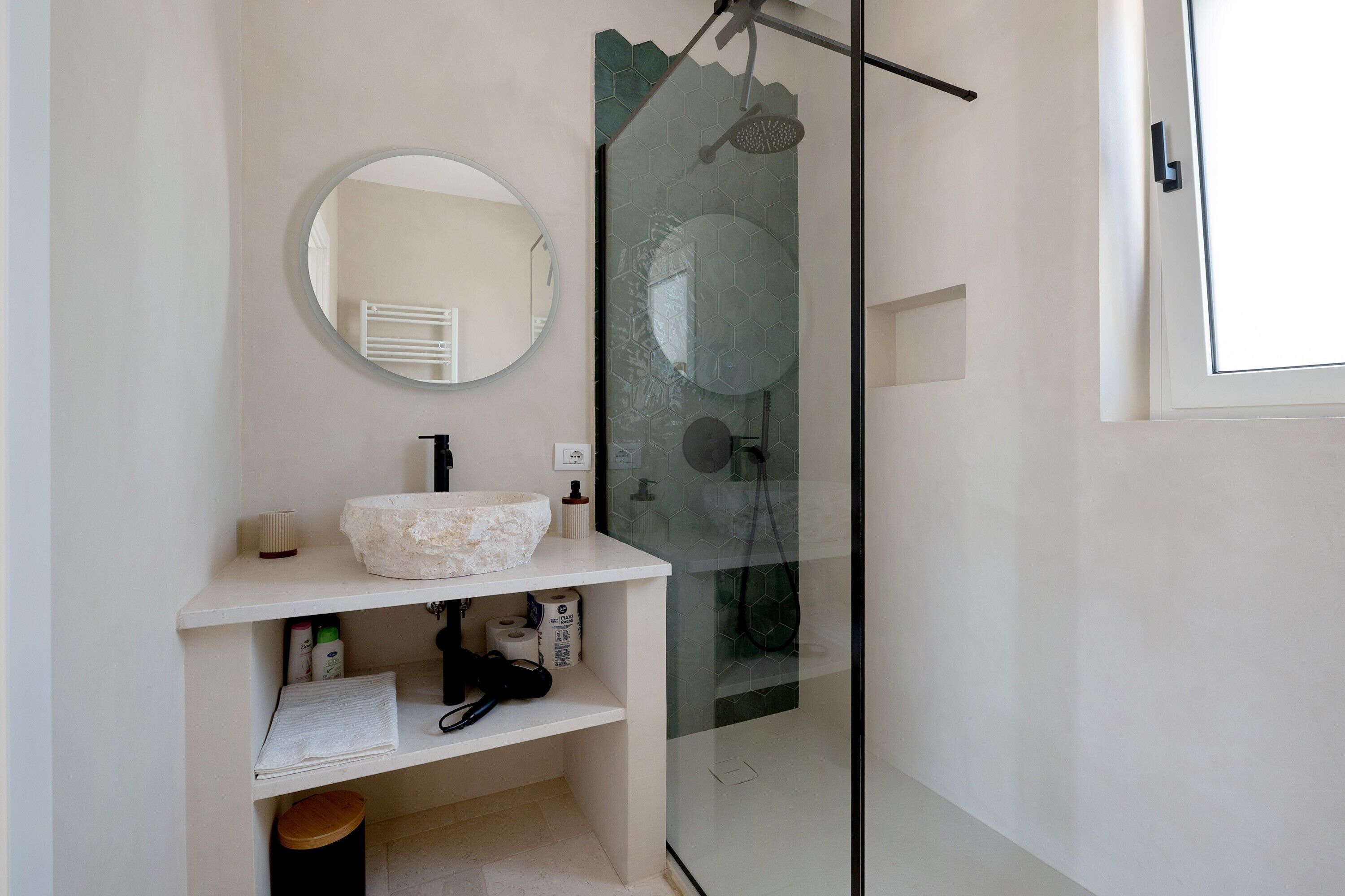 Une salle de bain avec un miroir rond, un lavabo en pierre, une douche avec une porte en verre et des toilettes.