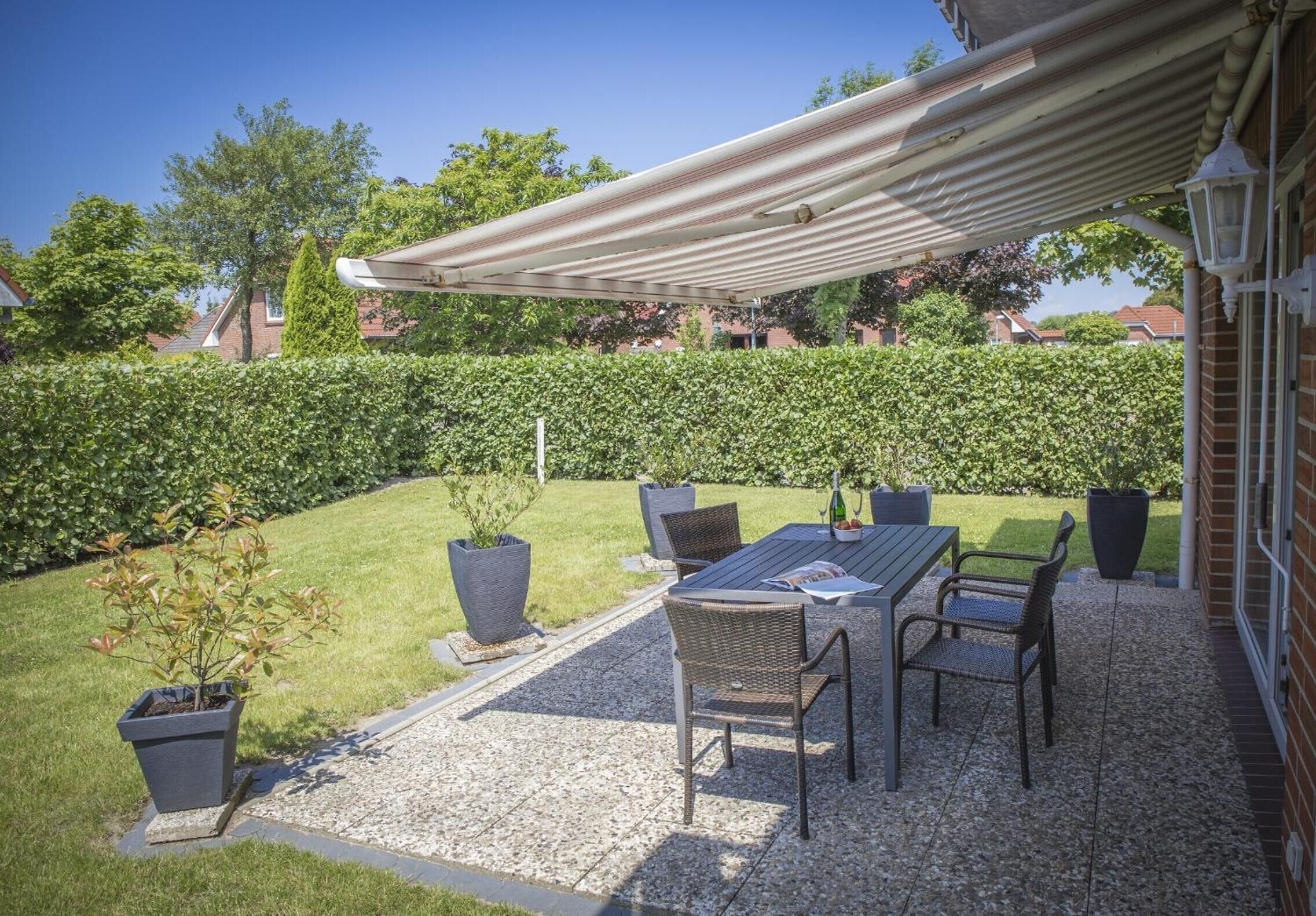 Eine Terrasse im Freien mit Tisch und Stühlen, umgeben von einer Hecke und einem Rasen.