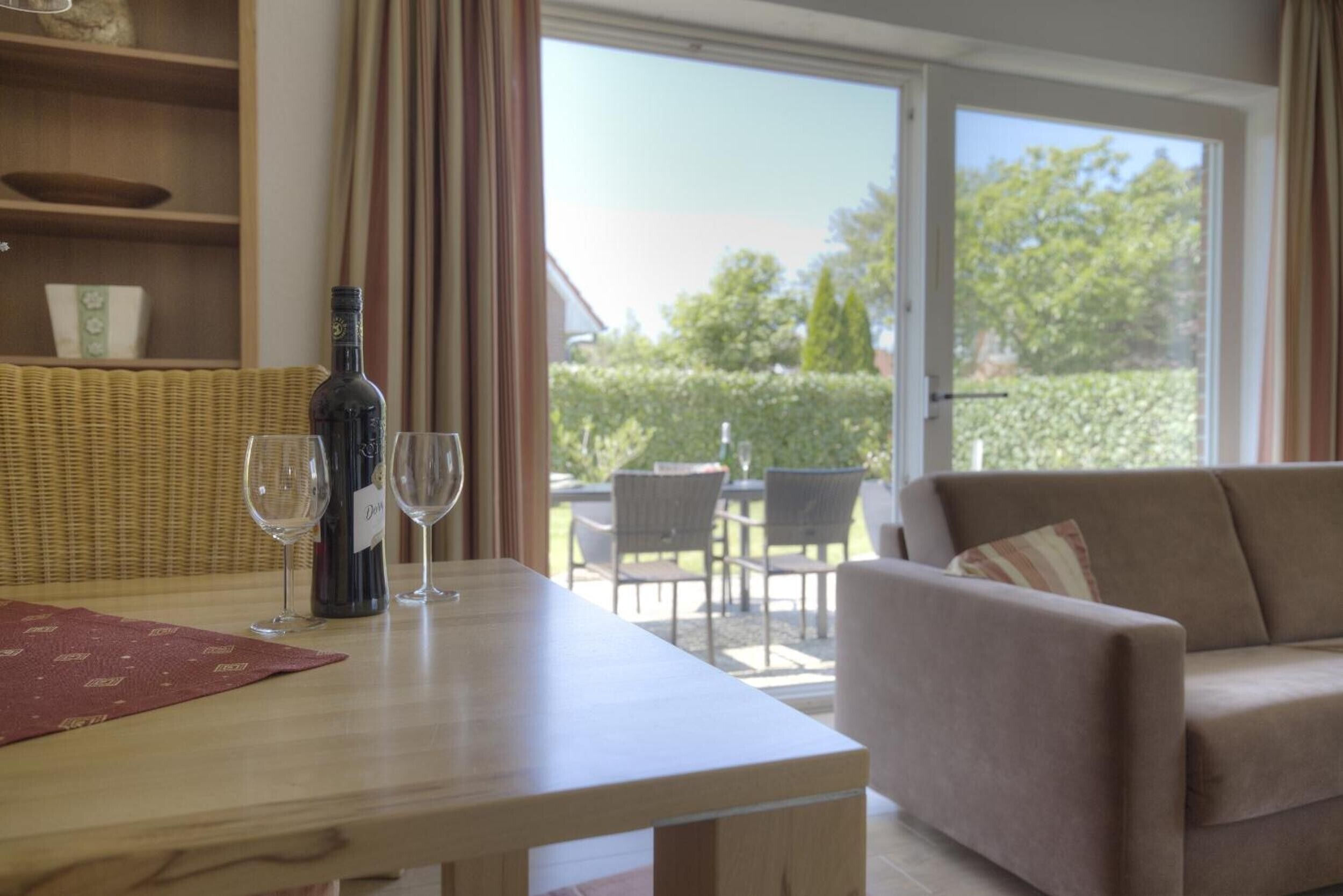 A room with a dining table, two wine glasses, a bottle, a sofa, and a view of the outdoors.