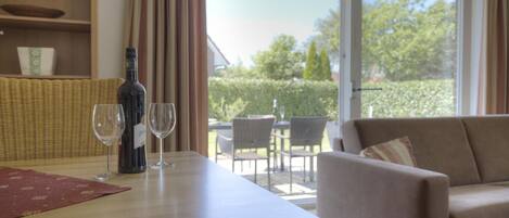 A room with a dining table, two wine glasses, a bottle, a sofa, and a view of the outdoors.