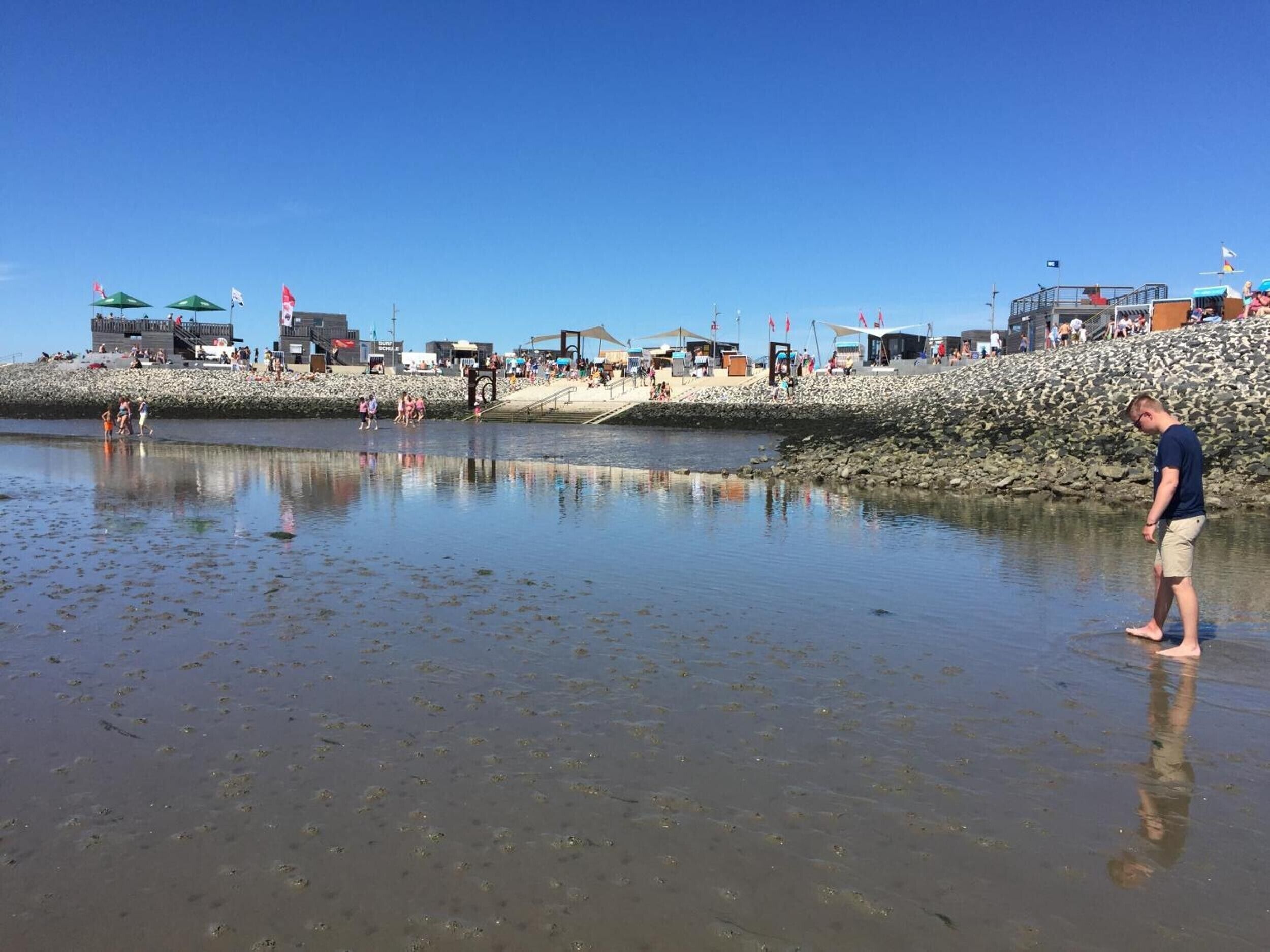 Una playa con costa rocosa, gente caminando y un hombre de pie en aguas poco profundas.