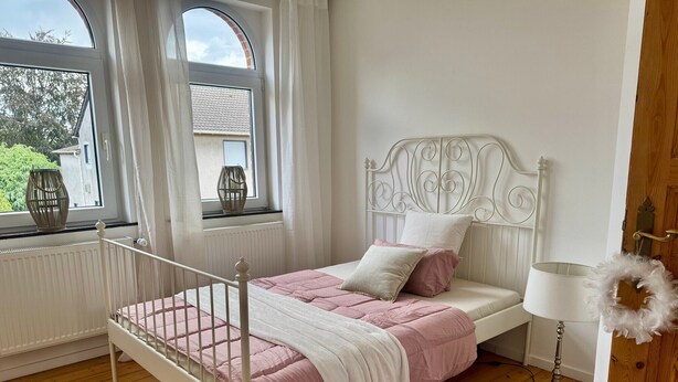 Quarto com cama, janela com cortinas, abajur e piso de madeira.