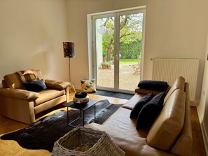 A living room with a sofa, a coffee table, and a floor lamp. There is a sliding glass door leading to an outdoor area with plants and a chair. - Holiday Home 'Haus um 1900 – Entire Place for You' with Terrace, Garden & Wi-Fi (Ennigerloh)