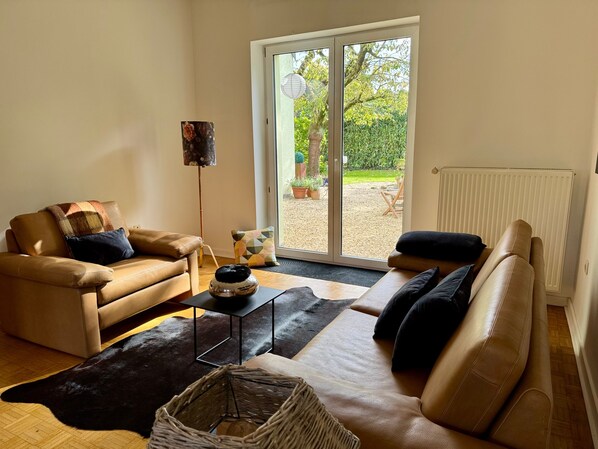 Ein Wohnzimmer mit einer Couch, einem Couchtisch und einer Stehlampe. Ein Schiebeglastür führt zu einem Außenbereich mit Pflanzen und einem Sessel.