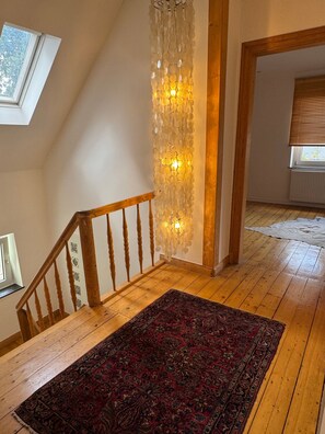 A wooden staircase with a handrail, a patterned rug on the floor, a wall-mounted light fixture, and a window with blinds. - Holiday Home 'Haus um 1900 – Entire Place for You' with Terrace, Garden & Wi-Fi (Ennigerloh)