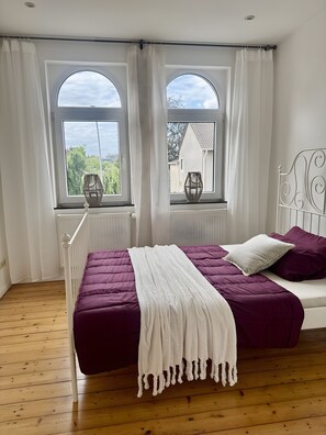 Ein Schlafzimmer mit einem Bett, zwei Vorhängen, einem Heizkörper und Blick nach draußen.