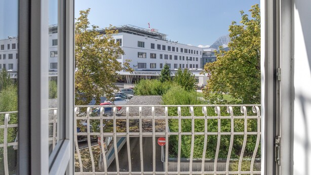 A view through a window with a metal railing, overlooking a parking area with cars and a building with multiple windows and a sign. - Apartment “Grattschlössl 1” with Mountain View, Balcony, and Wi-Fi (Sankt Johann)