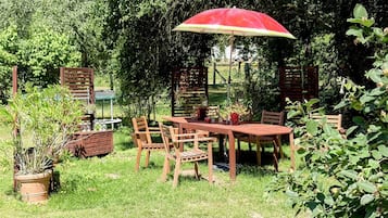 An outdoor patio with a wooden table and chairs, surrounded by greenery and a red umbrella.