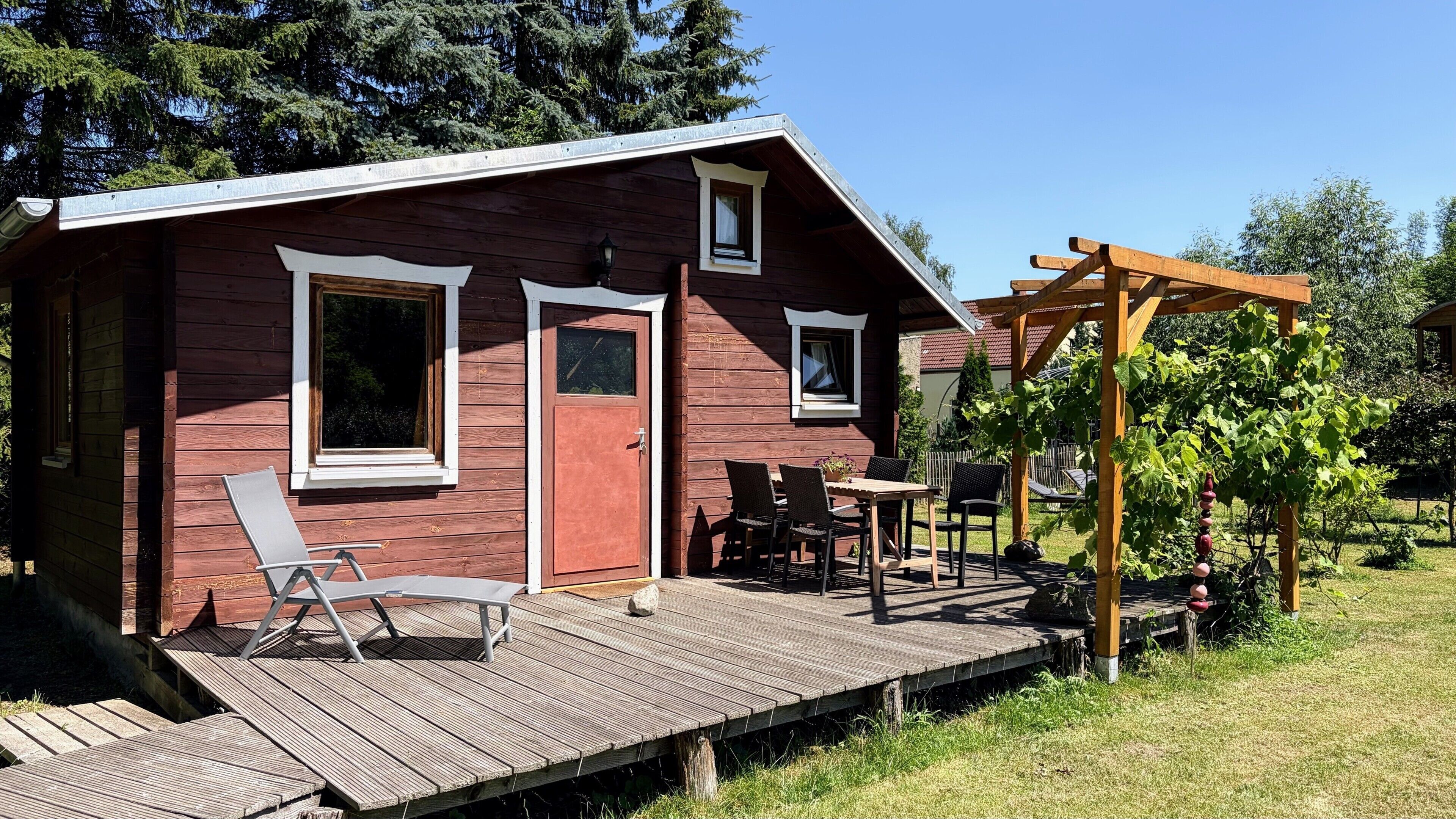A wooden cabin with a deck, two windows, and a pergola with vines.