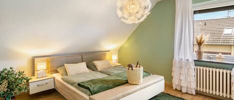 Un dormitorio con cama, mesitas de noche, lámpara colgante, ventana con cortinas y radiador.