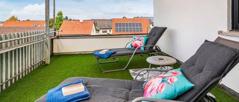 Een balkon met twee loungestoelen, waarvan er een een blauwe handdoek en een boek heeft, en de andere een roze kussen met bloemen.