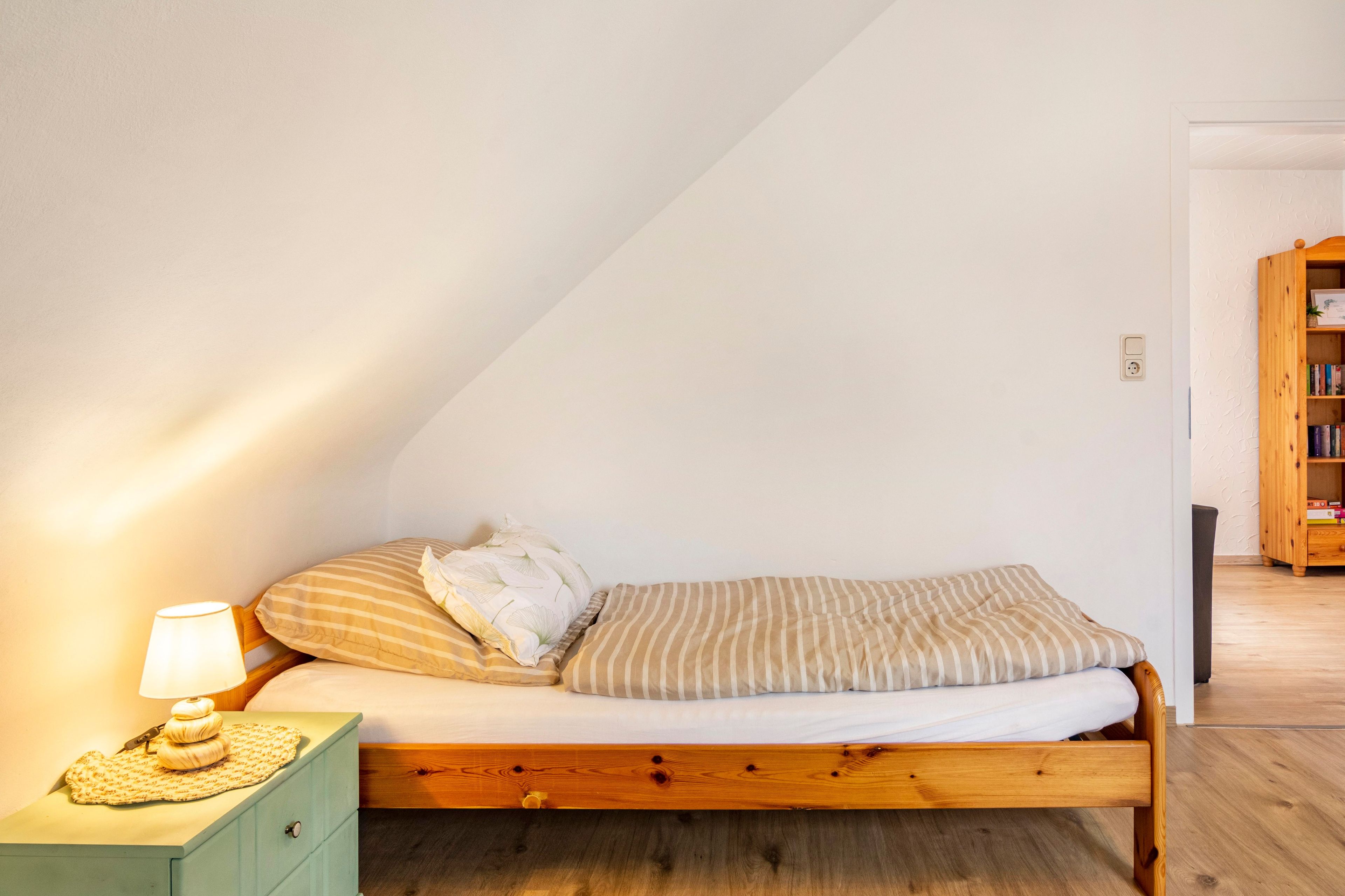 En enkeltseng med træramme, et sengebord med lampe og en bogreol i et rum med skrå vægge.