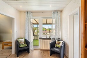 Een kamer met twee zwarte stoelen, een houten tafel, een radiator en uitzicht op een tuin door een raam met gordijnen.