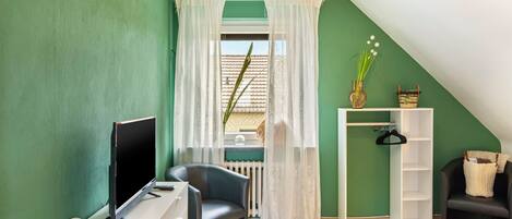 Quarto com teto inclinado, uma escrivaninha com um computador, uma cadeira, uma janela com cortinas brancas e uma pequena estante com objetos decorativos.